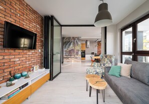 A modern living room with a brick wall, a flat-screen TV, a sofa, and a dining area with a table and chairs. - Metro Płocka Urban Oasis Apartment (Warszawa)