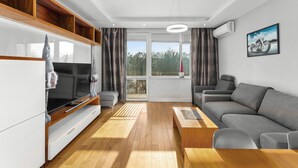 A modern living room with a sofa, a coffee table, a TV, and a balcony with a view of trees. - Nowa Huta Charming Apartment (Kraków)