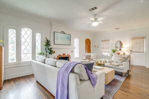 Ein Wohnzimmer mit einem weißen Sofa, einem Couchtisch und einem Kamin. Drei Fenster mit weißen Rahmen und ein Deckenventilator sind ebenfalls vorhanden.