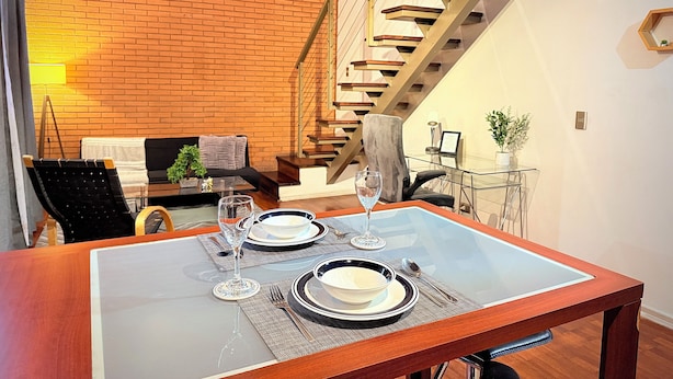 Un comedor con una mesa de vidrio dispuesta para dos personas, una escalera de madera y una pared de ladrillo al fondo.