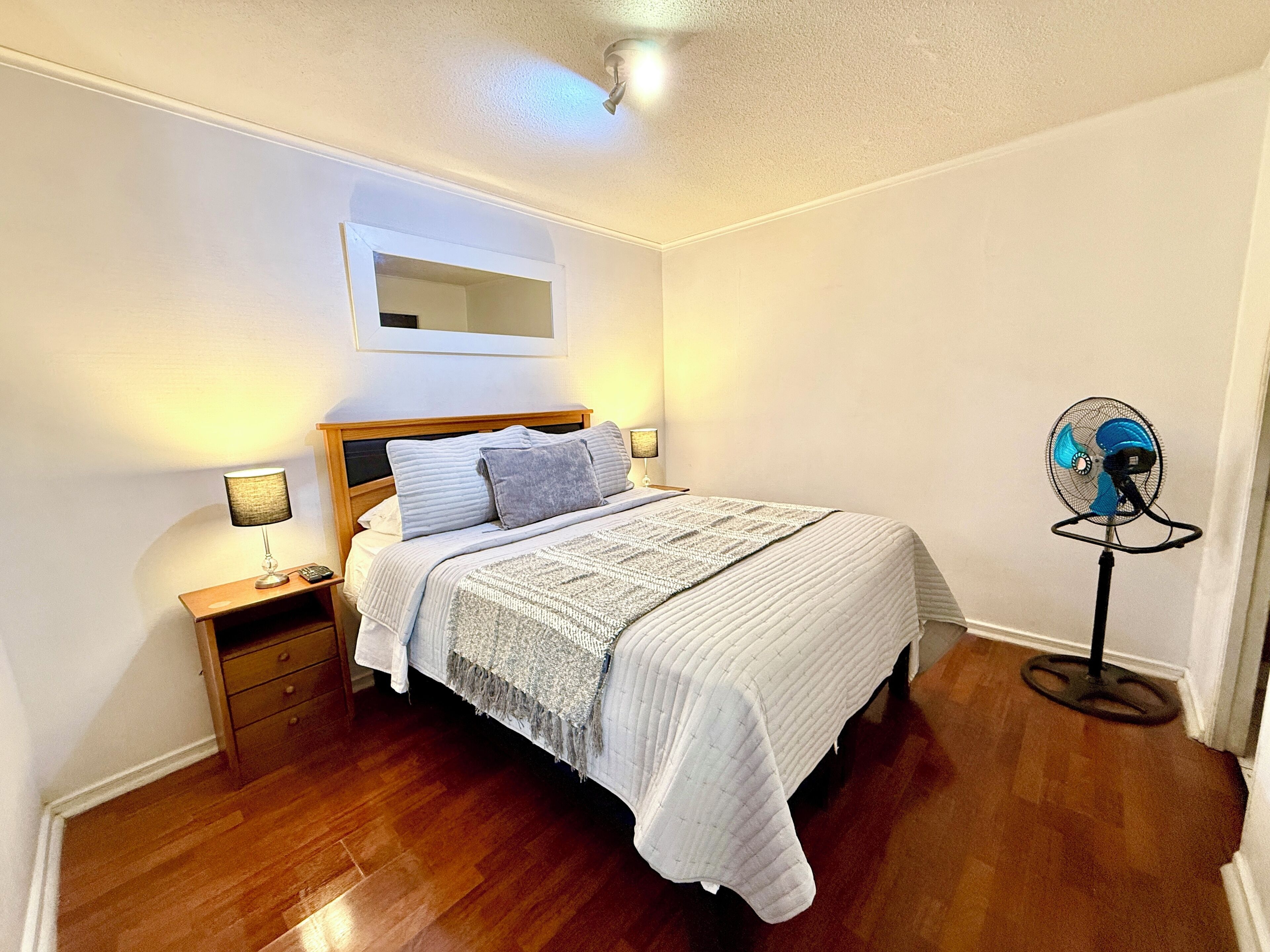 Une chambre avec un lit en bois, des lampes de chevet, un ventilateur sur pied et un miroir au mur.