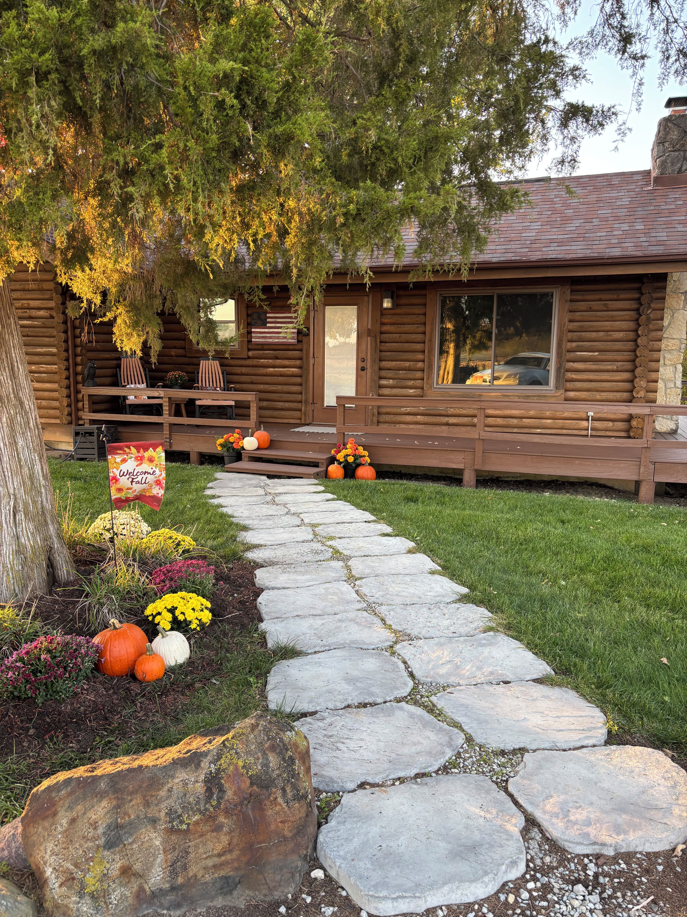 A wooden cabin with a stone pathway leading to the entrance, surrounded by autumnal foliage and decorative pumpkins.