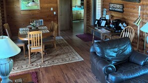 L’intérieur d’un chalet rustique comprend un coin repas, un canapé en cuir, des poutres en bois et une cuisine visible à travers une porte ouverte.