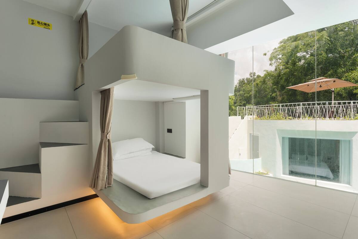 A modern bedroom with a built-in bed, a small seating area, and a balcony with a view of the trees.