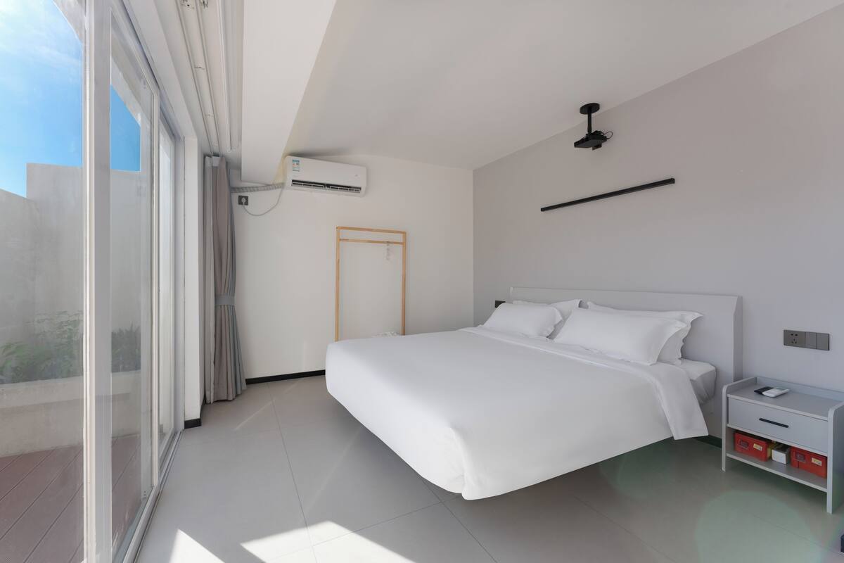 A modern bedroom with a large bed, a bedside table, a nightstand, a ceiling fan, an air conditioning unit, and a sliding glass door leading to an outdoor balcony.
