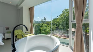 Een moderne badkamer met een groot bad, een groene stoel en uitzicht op een weelderige tuin door grote ramen.