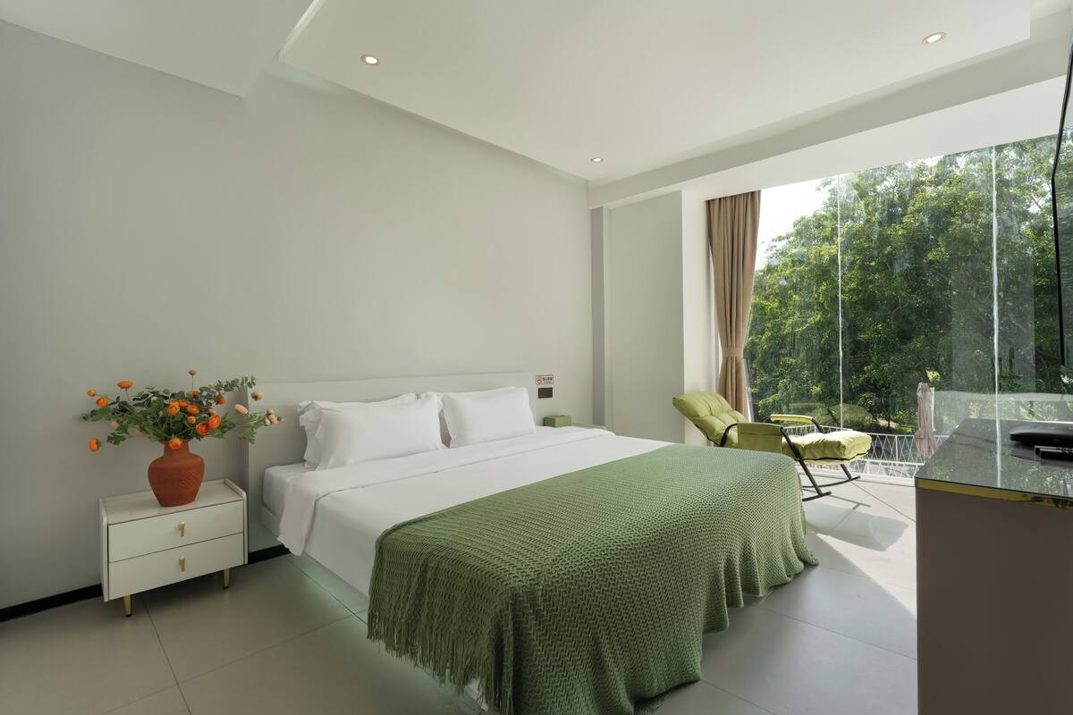 A modern bedroom with a large bed, a nightstand with a vase of flowers, a green blanket, and a balcony with a chair and table.