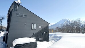 Un bâtiment moderne dont la façade porte l’inscription « IZUMIKO APARTMENT », entouré de neige et avec une montagne en arrière-plan.