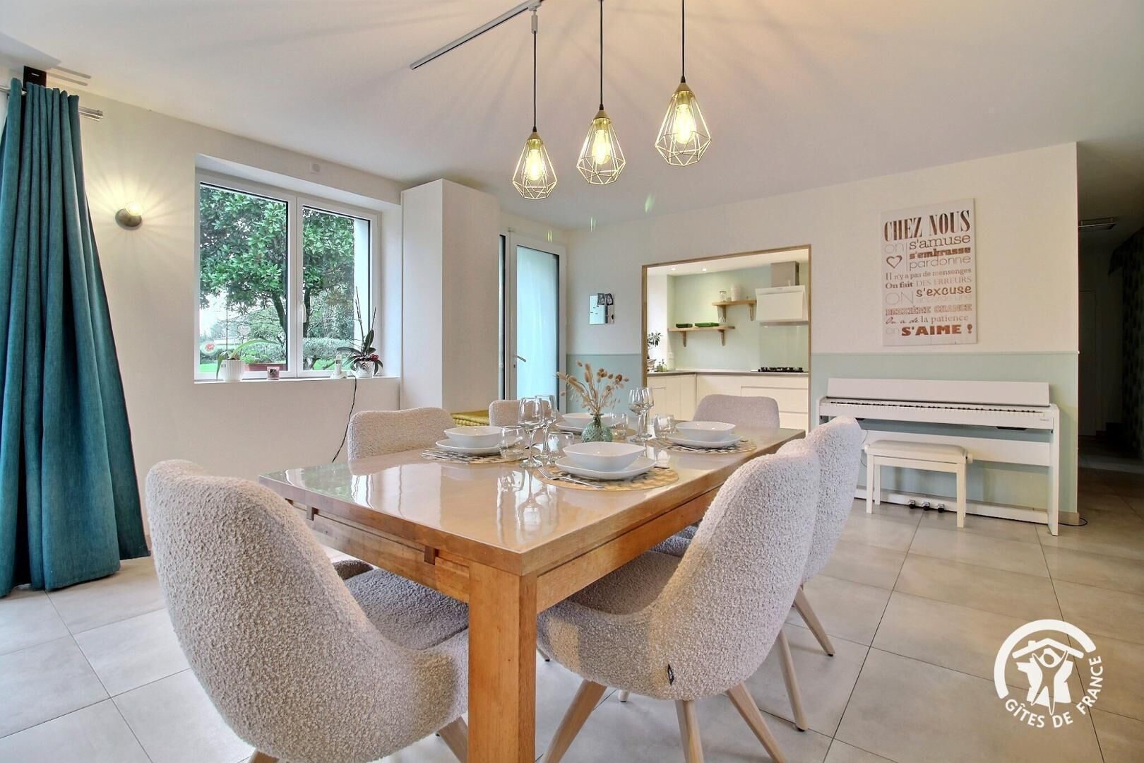 A dining area with a wooden table set for four, surrounded by upholstered chairs. There is a piano to the right and a large window with curtains on the left.