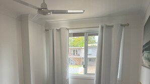 Ein Schlafzimmer mit Bett, Deckenventilator, Fenster mit Vorhängen und Blick auf ein Gebäude.