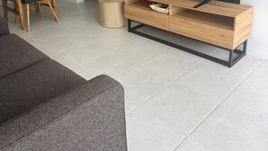 Ein modernes Wohnzimmer mit Flachbildfernseher auf Holzfuß, einem grauen Sofa, einem Essbereich mit Mikrowelle und einer kleinen Küche.