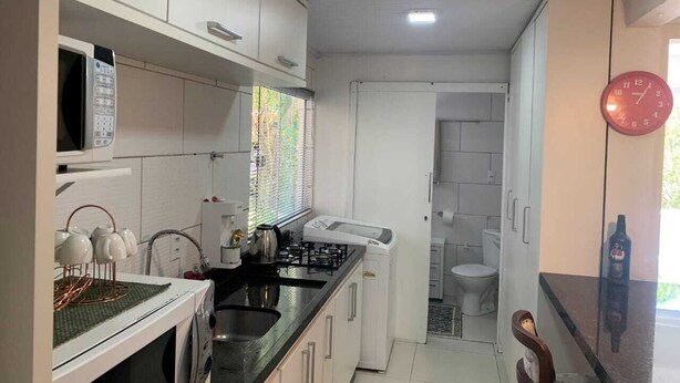 Cocina compacta con armarios blancos, un microondas, una estufa a gas y un lavabo. Hay una ventana con persianas, un reloj en la pared y una botella sobre la encimera.