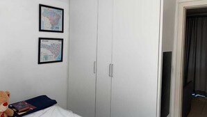 Un dormitorio con un armario blanco, dos mapas enmarcados en la pared, una cama con un cojín azul y un oso de peluche.