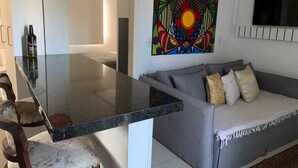 Una sala de estar moderna con una mesa de comedor de vidrio, un sofá gris con cojines, un cuadro abstracto colorido y un reloj rojo en la pared.