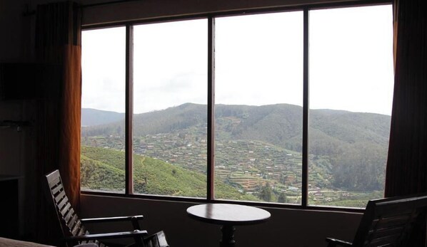 Habitación con un ventanal grande que ofrece vistas a un valle y montañas. En primer plano, se aprecia una mesa redonda y dos sillas.