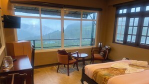 A hotel room with a bed, a desk, a chair, a small table, and a window with a view of a mountainous landscape. - House of Leisure Holiday Bungalow  (Nuwara Eliya)