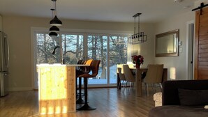 Een ruime woonkamer met houten vloer, een donkergrijze bank, een eettafel met stoelen, en een keukenhoek met houten aanrecht en barstoelen.