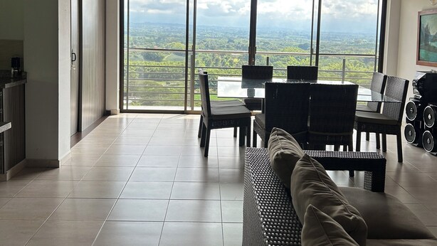 A spacious living area with a high ceiling, large windows offering a view of the countryside, a dining table with chairs, and a sofa.
