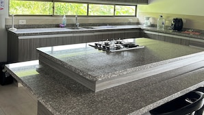 A modern kitchen with a large island, granite countertops, and stainless steel appliances. There is a window providing natural light and a view of greenery outside.