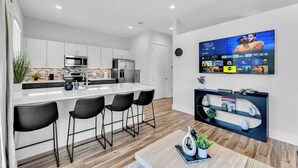A modern kitchen with a central island, black bar stools, a flat-screen TV mounted on the wall displaying a streaming service, and a shelf with books and decorative items. - NEW Modern 1BR in Oak Lawn Dallas With Free Parking + Complimentary EV Charger (Dallas)