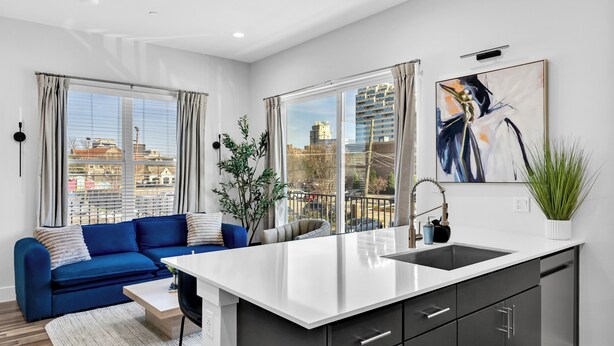 A modern apartment with a kitchen, dining area, and living room. It features large windows, a blue sofa, and a modern kitchen with a white countertop. - NEW Modern 1BR in Oak Lawn Dallas With Free Parking + Complimentary EV Charger (Dallas)