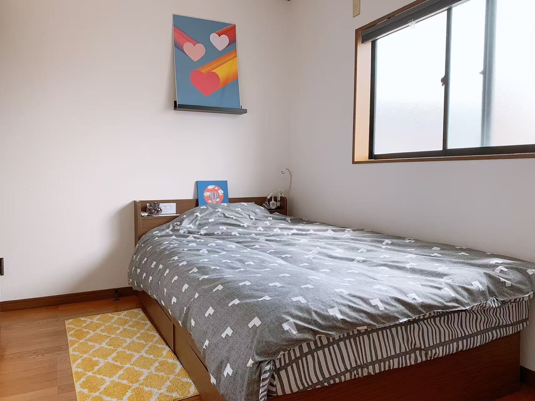 A single bed with a patterned blanket, a bedside table with a lamp, a wall clock, and a window with blinds.