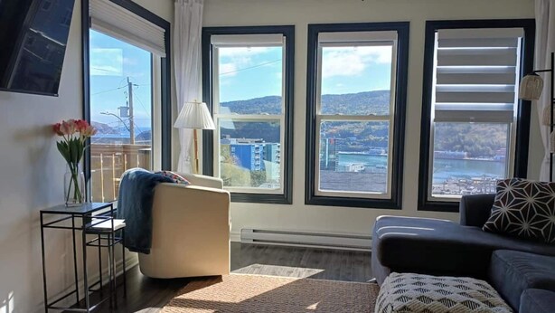A modern living room with large windows, a sofa, a chair, and a small table with a vase of flowers. - Lofty Haven - High/ Harbour Views of Narrows & Signal Hill/ Steps to Downtown (St. John's)
