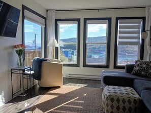 A modern living room with large windows, a sofa, a chair, and a small table with a vase of flowers. - Lofty Haven - High/ Harbour Views of Narrows & Signal Hill/ Steps to Downtown (St. John's)