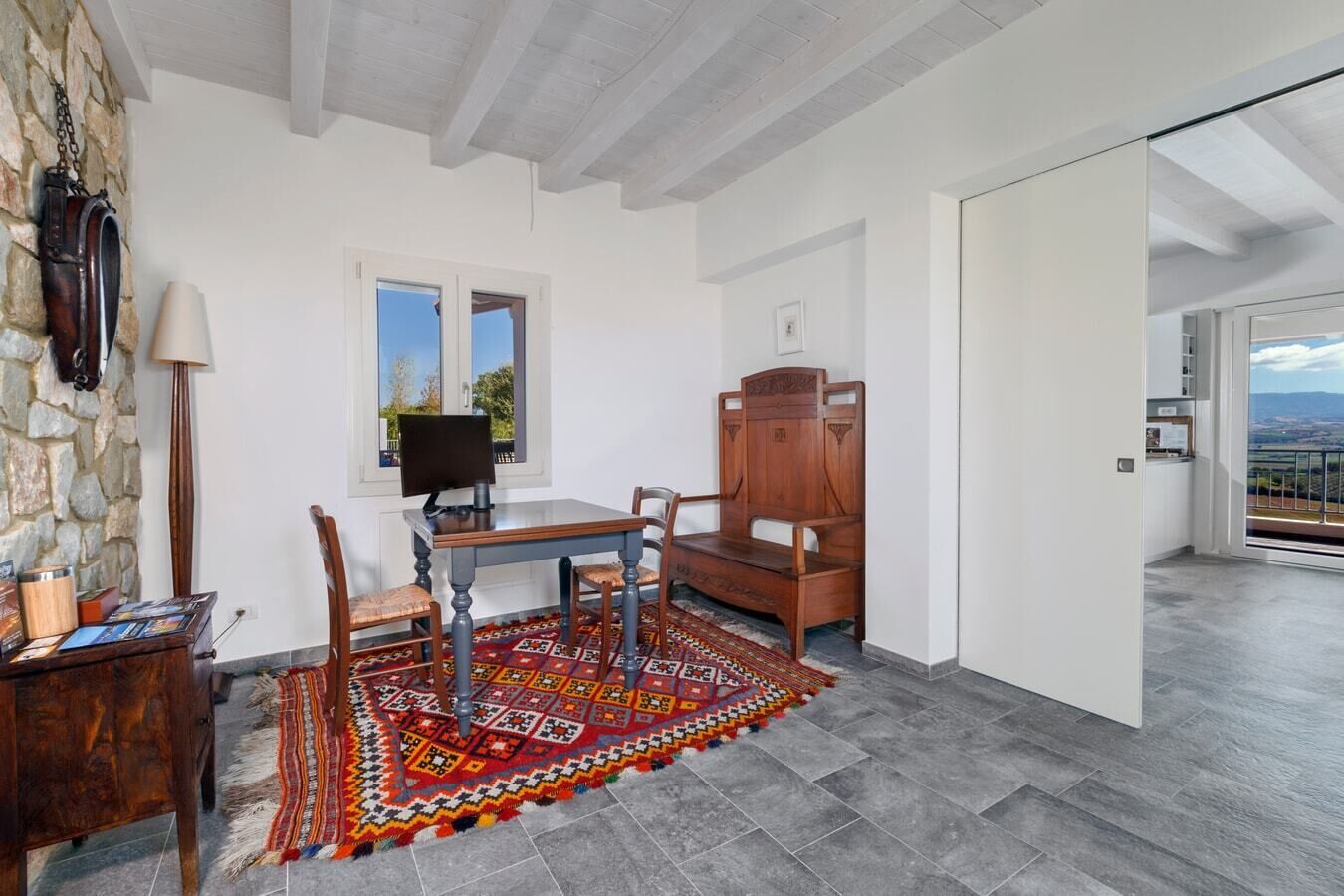 Une pièce avec un mur en pierre, une table en bois avec des chaises, un tapis à motifs et une vue sur l’extérieur, visible à travers une porte vitrée.