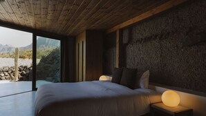 Een slaapkamer met een groot bed, houten plafond, stenen muur en uitzicht op bergen en cactussen.