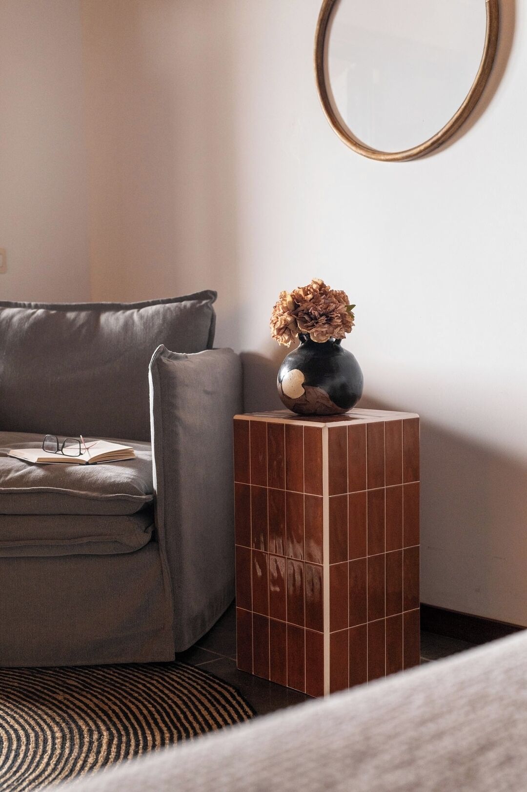 Een hoek van een kamer met een grijze bank, een klein zwart vaasje met gedroogde bloemen, en een ronde spiegel aan de muur.