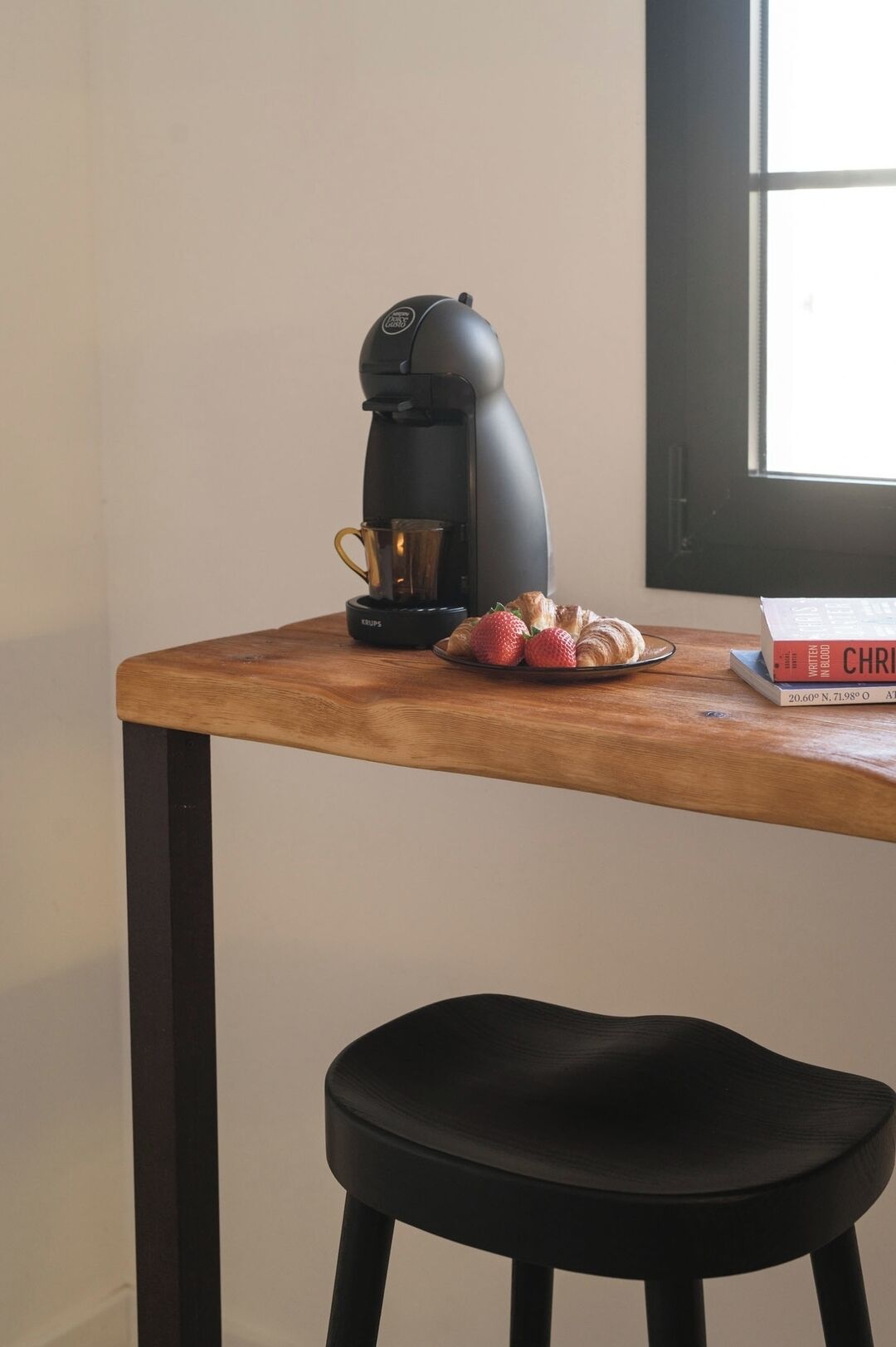 A wooden table with a black coffee maker, a cup, and a plate of strawberries and croissants. A black stool is in front of the table.
