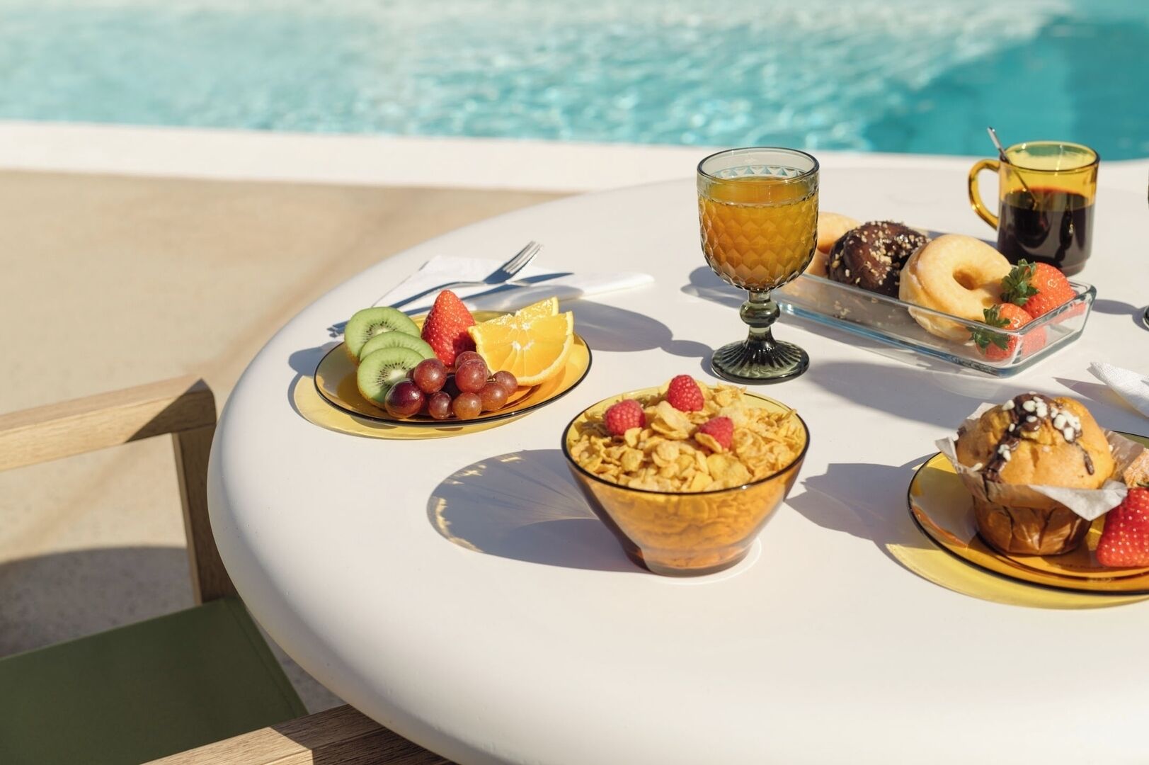 A table with breakfast items including fruits, cereal, a muffin, and a glass of juice by a pool.