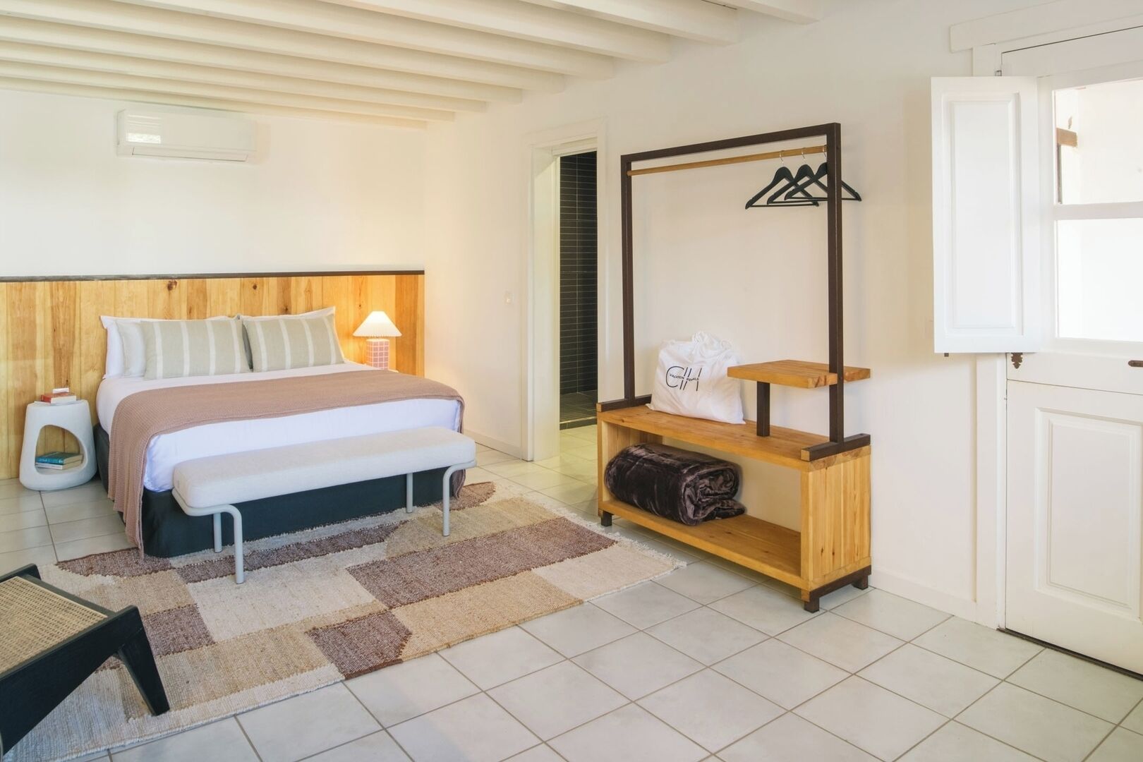 Een slaapkamer met een bed, nachtkastje, lamp en een houten plank met een tas.