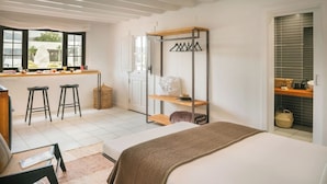 Une chambre avec un lit, un bureau avec deux tabourets, un portant à vêtements et une vue sur l’extérieur grâce à de grandes fenêtres.