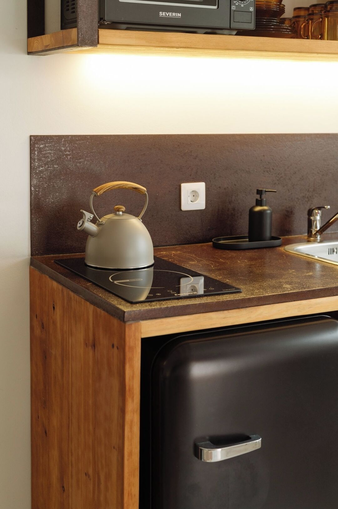 Una cocina compacta con tetera sobre una placa de inducción, un refrigerador negro y una encimera de madera.