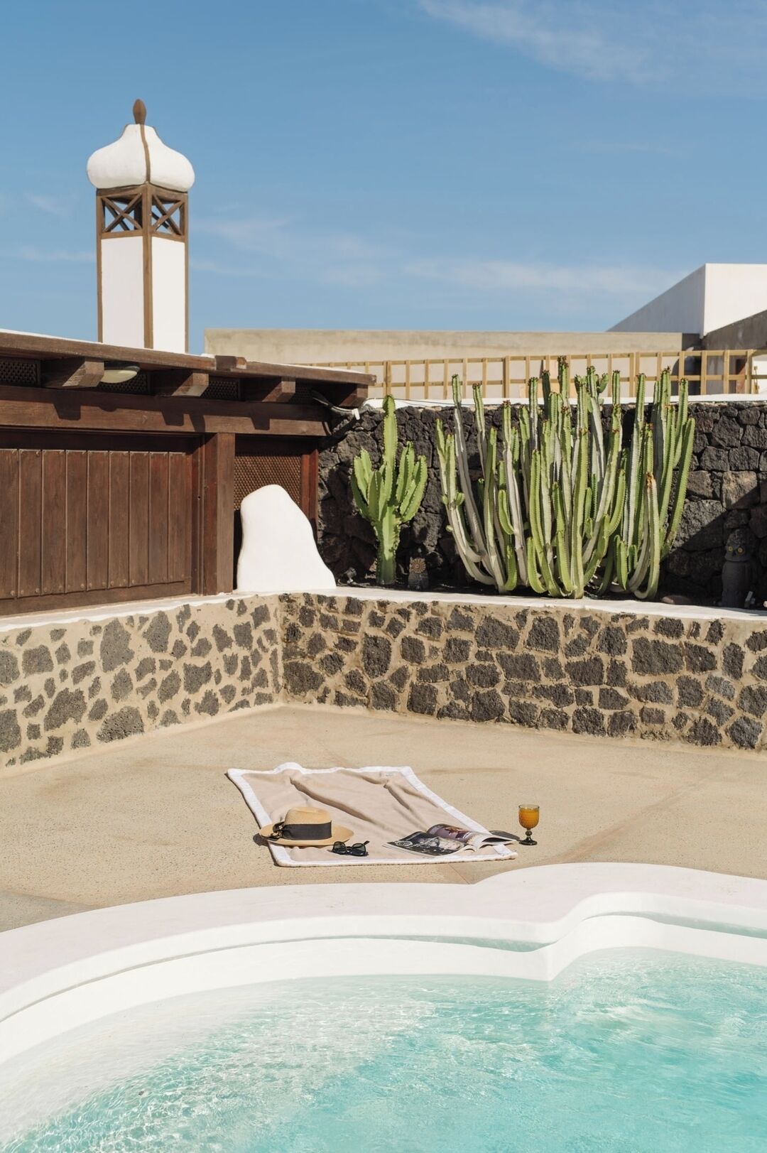 A hot tub area with a stone wall, a wooden pergola, and cacti.