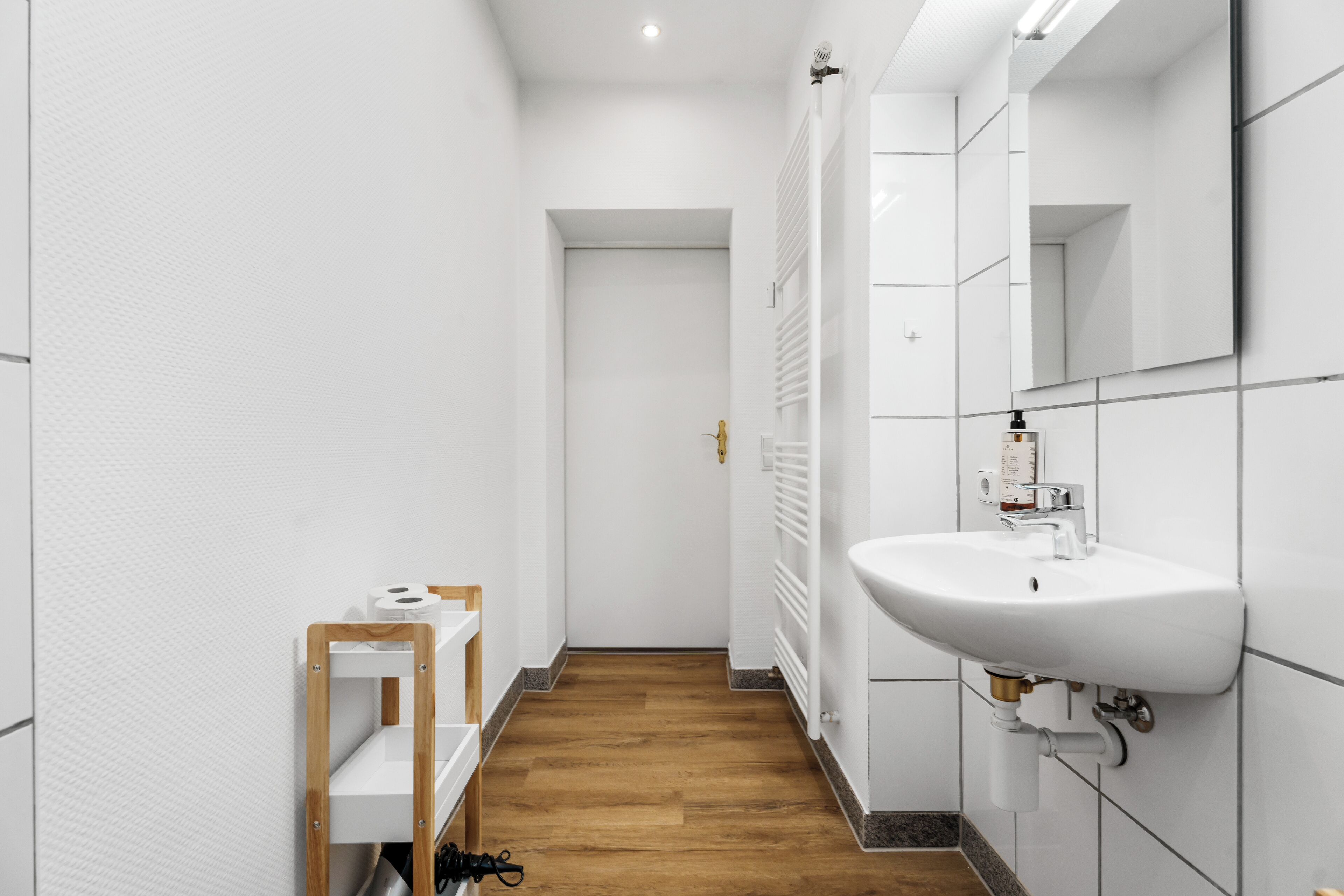 A narrow hallway with a white tiled wall, a wooden floor, a white sink with a mirror, and a radiator.