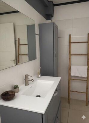 A bathroom with a white sink, a mirror, a grey cabinet, and a bamboo shelf with a towel. - Villa Tropicale - With Wellness Treatments (Complimentary) (Sainte-Anne)
