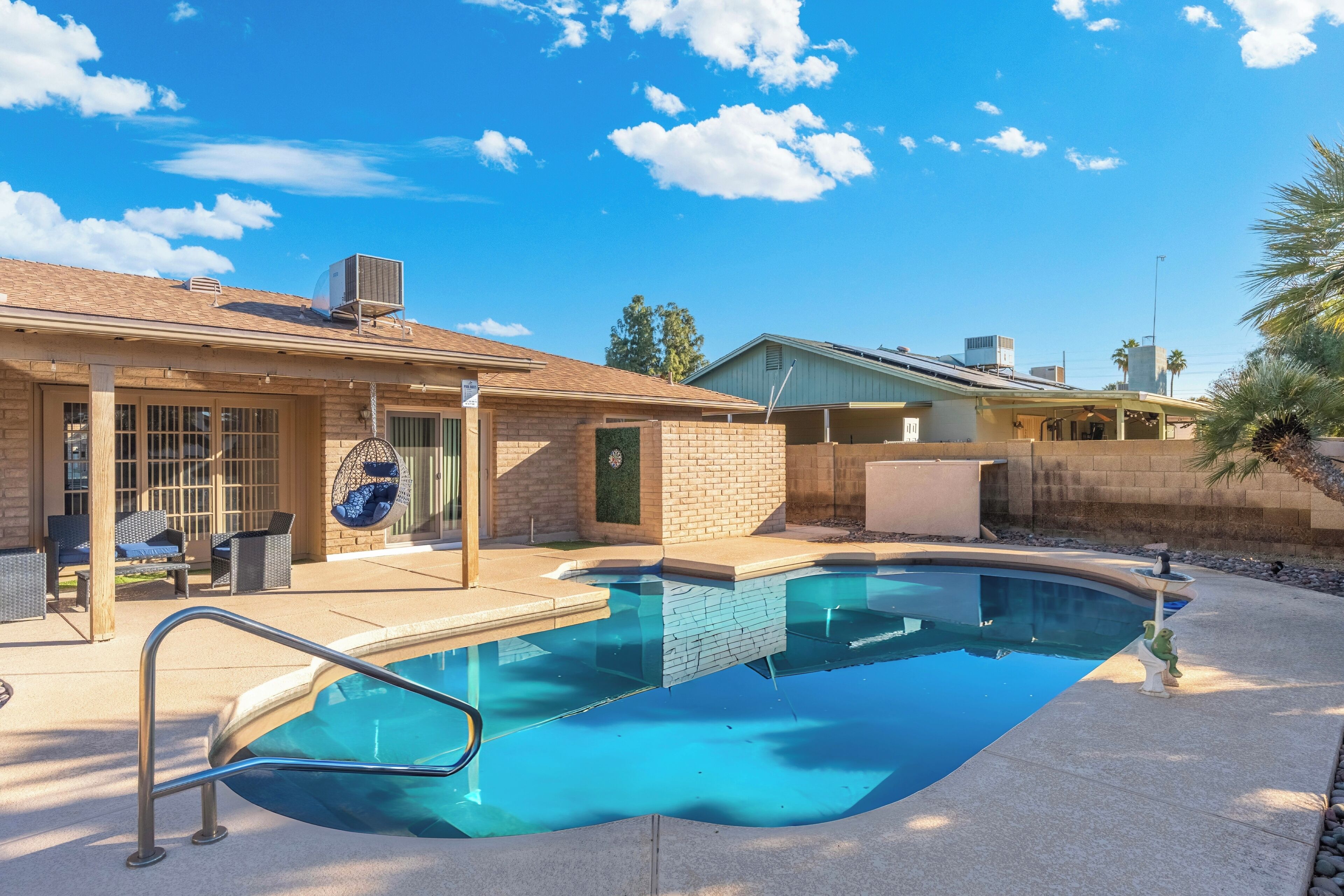 A swimming pool with a clear blue water, surrounded by a patio area with outdoor furniture and a brick building in the background.