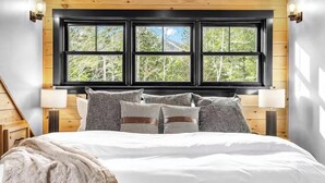 Een slaapkamer met een groot bed, meerdere kussens, een raam met uitzicht op bomen en houten lambrisering.