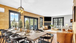Een ruime woonkamer met een grote eettafel, zwarte stoelen, een open haard en een aan de muur bevestigde televisie.