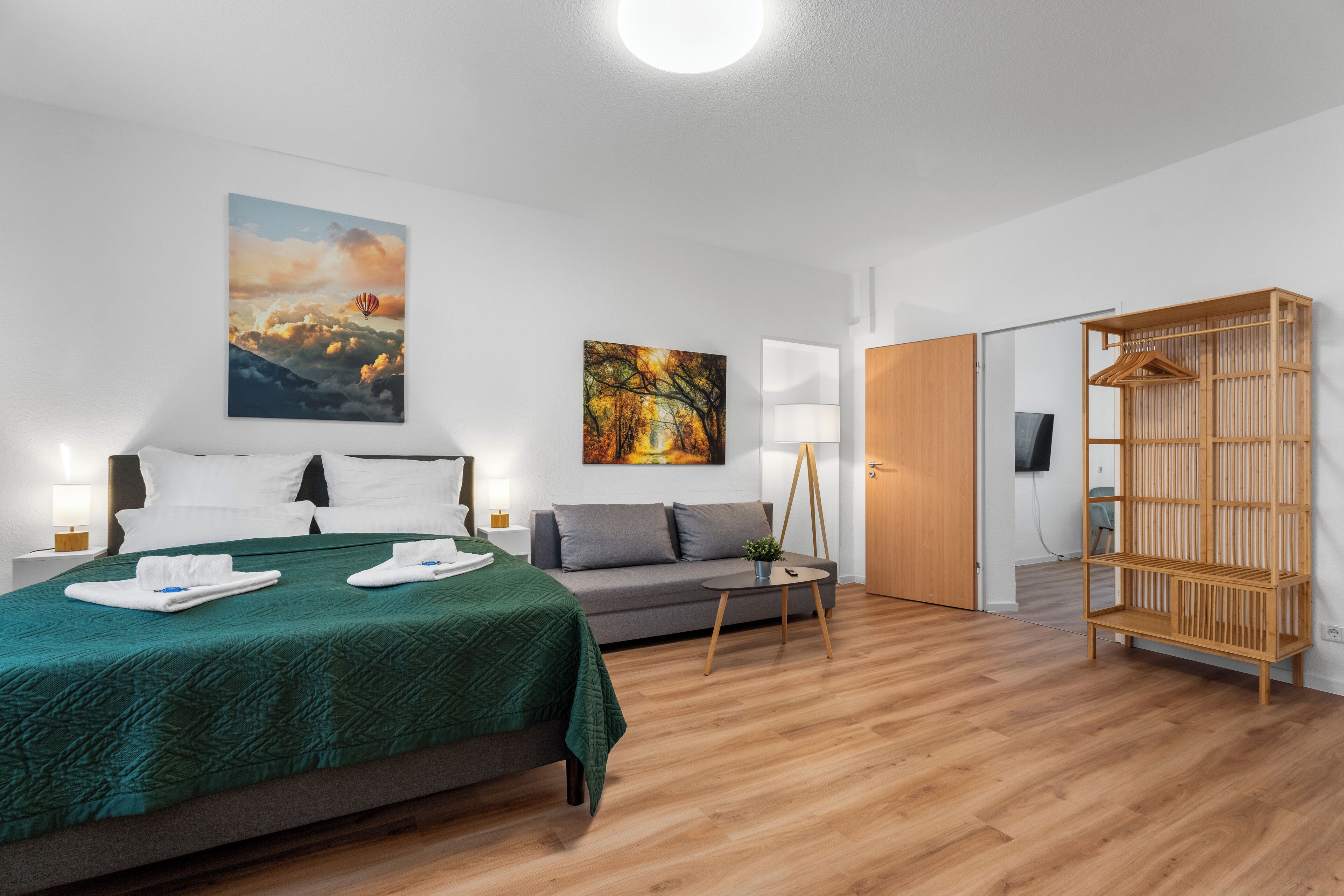 A modern hotel room with a bed, a sofa, a small table, a lamp, and a wooden wardrobe.