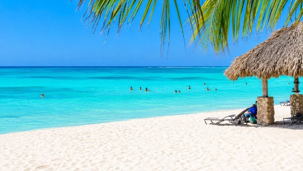 A beach with clear turquoise water, people swimming, and a thatched-roof structure with chairs. - Gold Coast Aruba  (Noord)