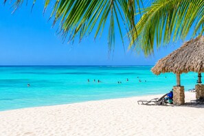 A beach with clear turquoise water, people swimming, and a thatched-roof structure with chairs. - Gold Coast Aruba  (Noord)