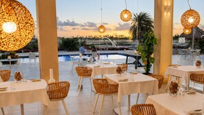 A restaurant with outdoor seating, white tables, wicker chairs, hanging lights, and a view of a pool and sunset. - Gold Coast Aruba  (Noord)