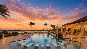 A swimming pool with lounge chairs, palm trees, and a view of the ocean at sunset. - Gold Coast Aruba  (Noord)