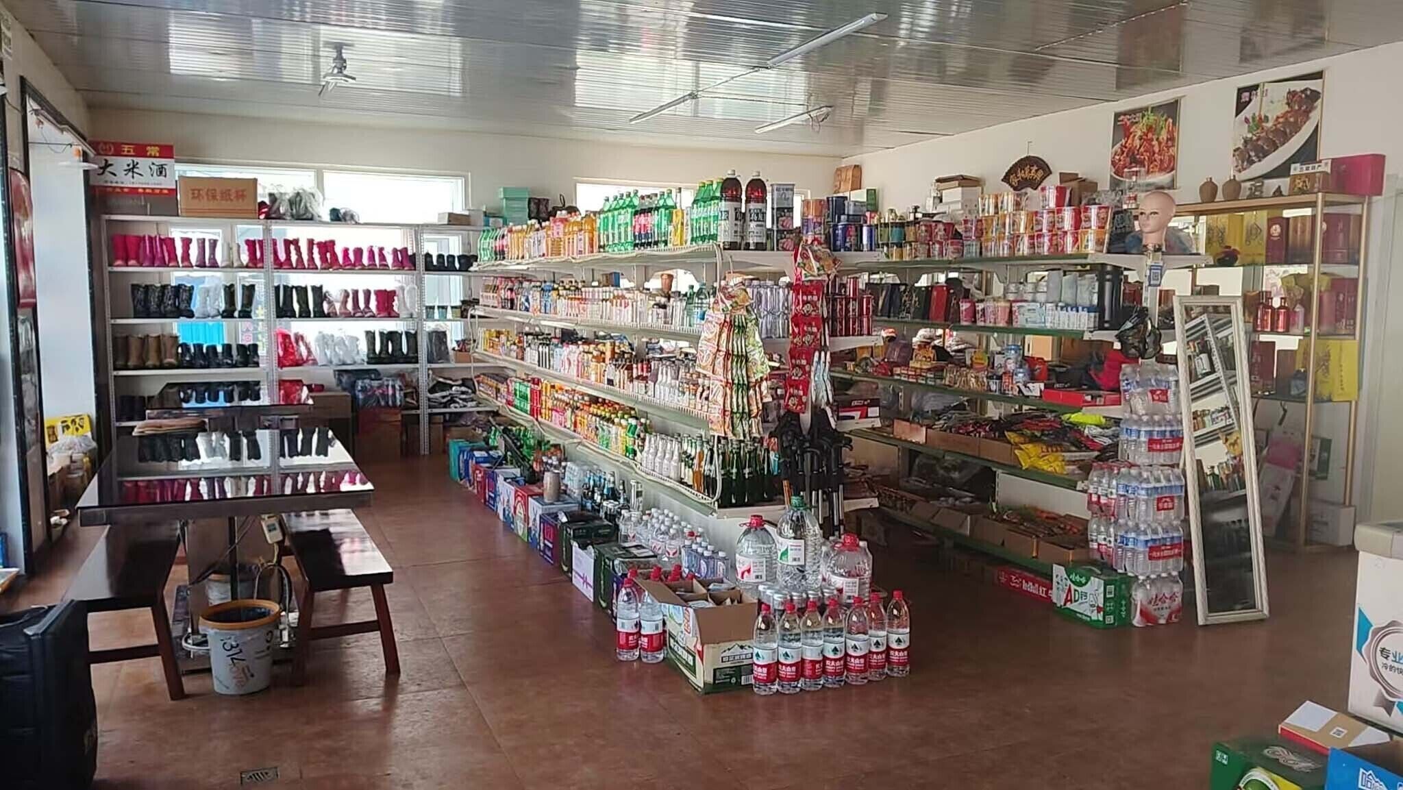 Une petite épicerie de quartier, avec des étagères remplies de divers produits, un comptoir avec une caisse enregistreuse et un présentoir de bouteilles d’eau.