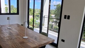 Une chambre moderne avec une table en bois, deux chaises, une lampe et une grande porte-fenêtre coulissante donnant sur un balcon avec vue sur la verdure et les bâtiments.
