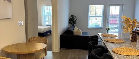 A modern kitchen with a central island, dining area with round tables, and a living space with a sofa and a plant.